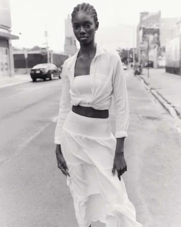 Black and white editorial portrait of a woman standing on a city street wearing a light-coloured skirt and knotted shirt.