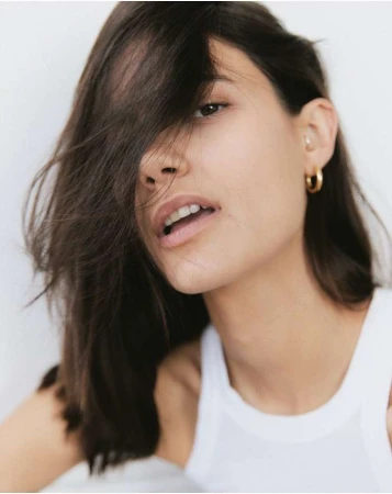 Close-up portrait of a woman with dark hair covering her eye, wearing a white tank top and gold hoop earrings against a white background.