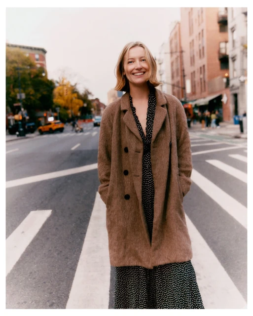 Woman in a brown coat stands on a crosswalk in an urban environment with brick buildings and cars visible in the background.