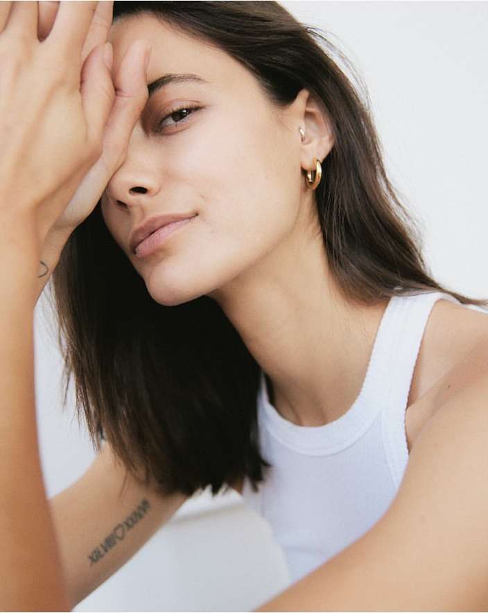 Close-up portrait of a woman wearing a white tank top and gold hoop earrings against a white background by Felix Wong.