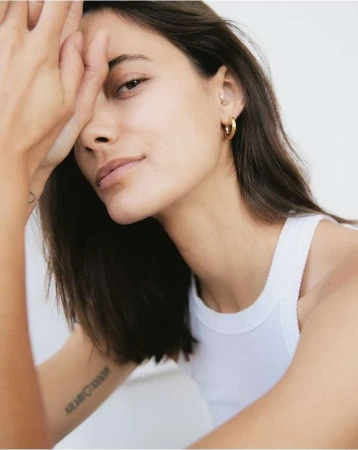 Close-up portrait of a woman wearing a white tank top and gold hoop earrings against a white background by Felix Wong.