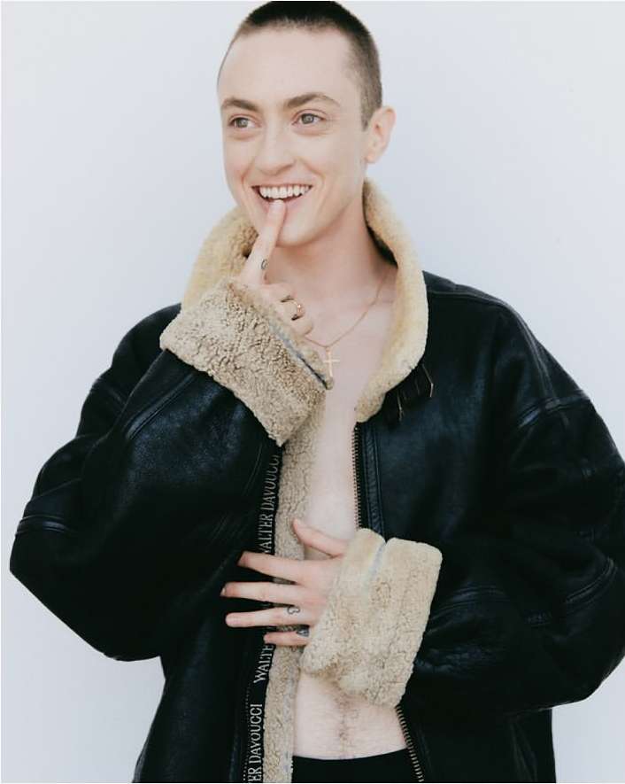 Portrait of a smiling adult with buzzed hair, posing in an open black jacket with a light tan fur lining and white studio backdrop.