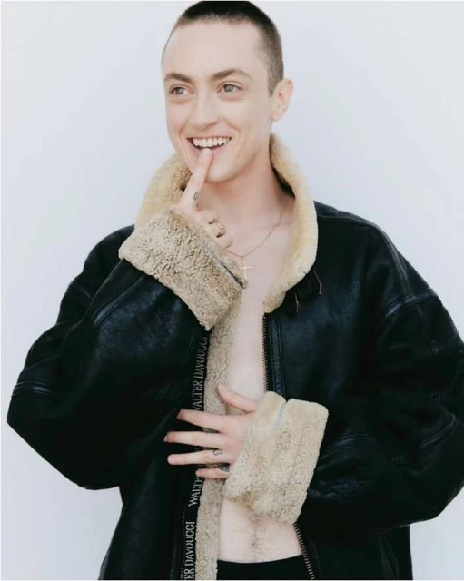 Portrait of a smiling adult with buzzed hair, posing in an open black jacket with a light tan fur lining and white studio backdrop.