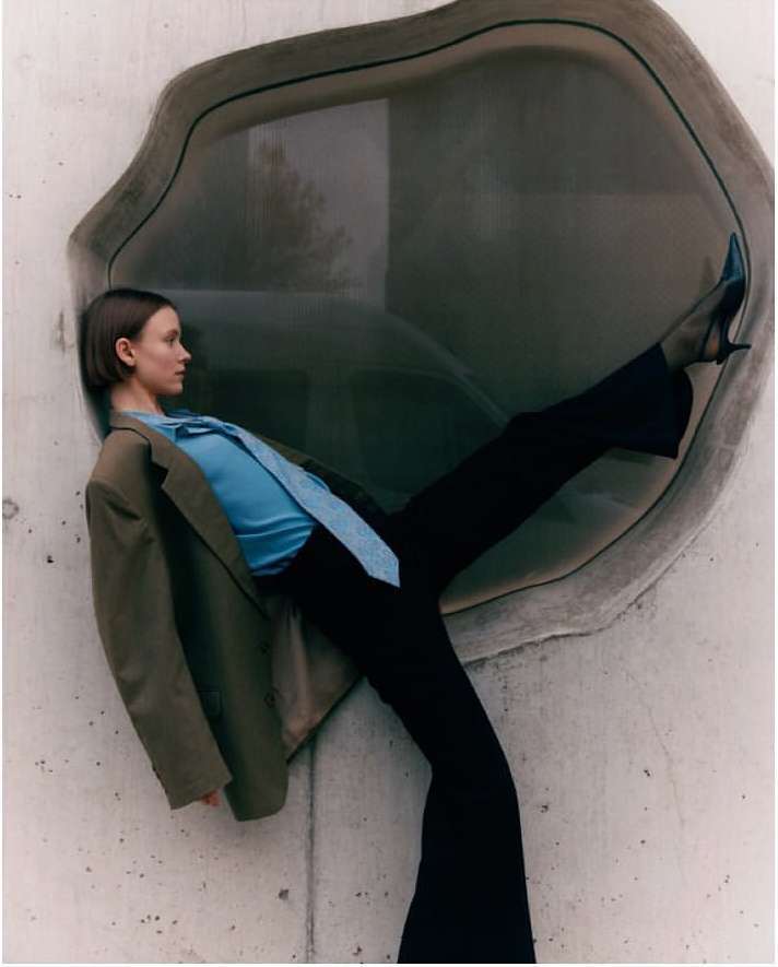 Fashionable woman poses in a teal blouse, wide-leg pants, and a tan blazer while leaning against a contemporary concrete wall with a window.