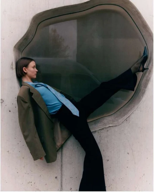 Fashionable woman poses in a teal blouse, wide-leg pants, and a tan blazer while leaning against a contemporary concrete wall with a window.