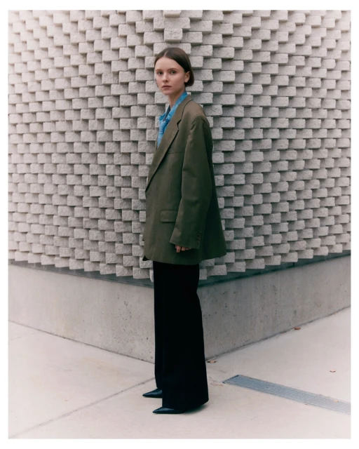 A young woman poses in an oversized olive jacket and black pants in front of a textured gray wall on a sunny day. Fashion editorial.