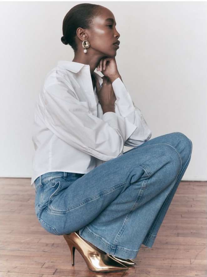 Elegant African-American woman in a white shirt and blue jeans posing indoors with gold high heels and wearing gold earrings.