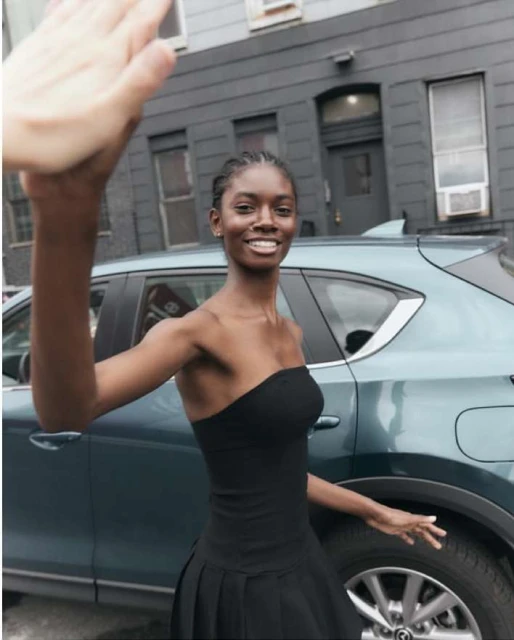 A smiling, attractive African-American woman in a black dress stands next to a modern teal SUV in front of a dark gray building.