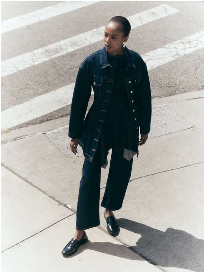 Woman in denim jacket and wide-leg jeans poses on crosswalk in natural light. She wears dark loafers, and shadows mark the street.
