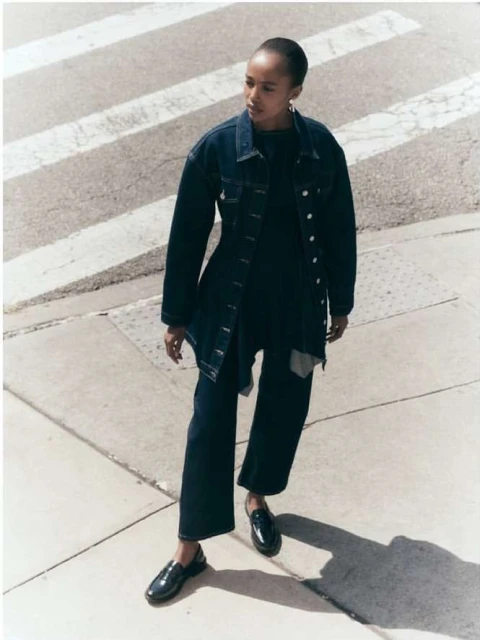 Woman in denim jacket and wide-leg jeans poses on crosswalk in natural light. She wears dark loafers, and shadows mark the street.
