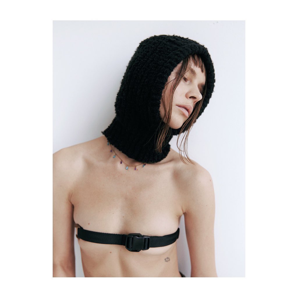 Close-up studio shot of an adult woman wearing a black knit balaclava and black strap across her bare chest against a white wall.