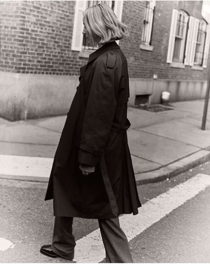 Woman in a long dark trench coat walking across a city street, captured in black and white photography by Felix Wong.