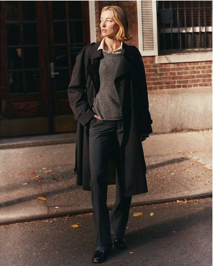 A female model in a black trench coat and grey sweater standing on an urban street by a brick building.