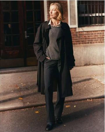 A female model in a black trench coat and grey sweater standing on an urban street by a brick building.