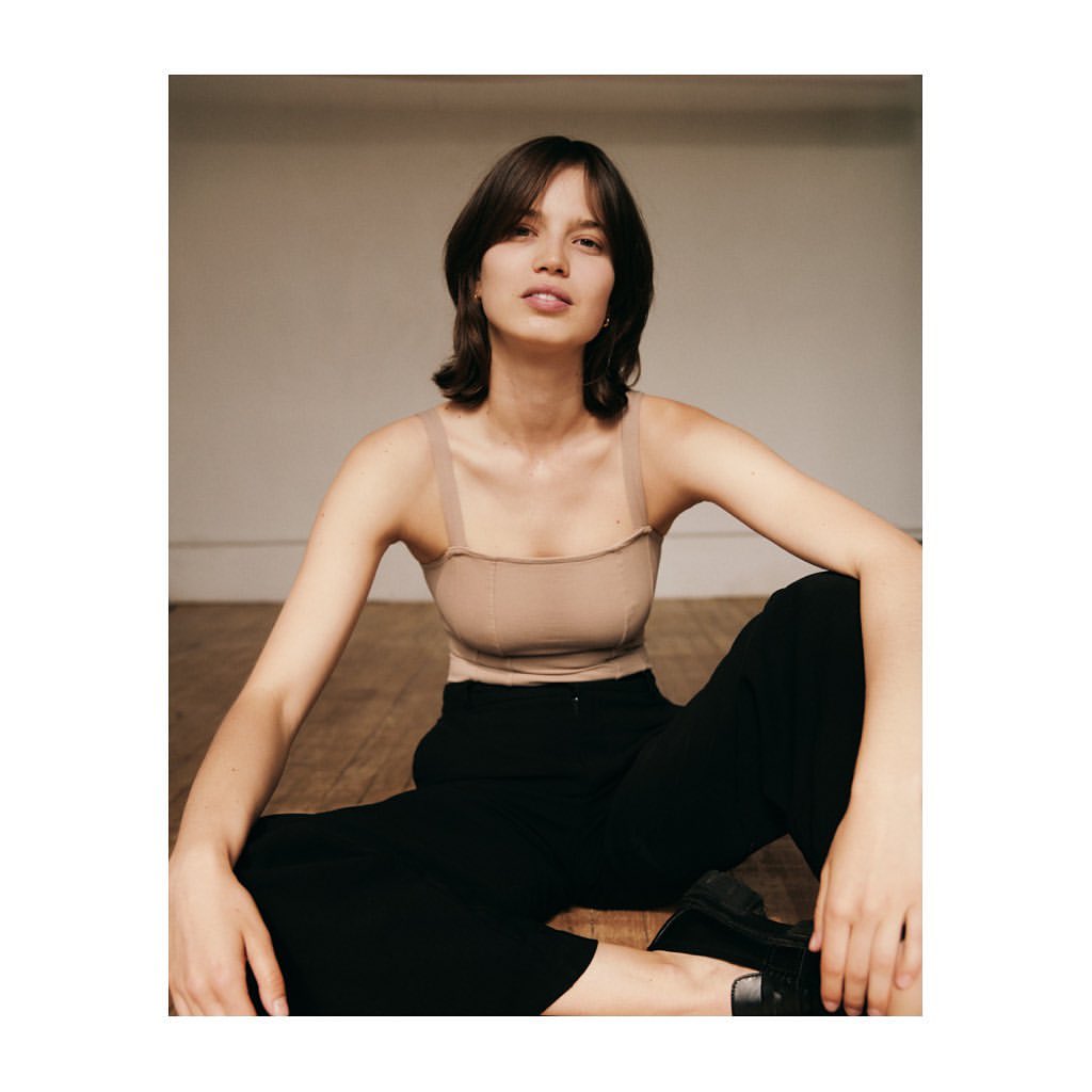 A young adult woman with short brunette hair sits on a wooden floor in a beige top and black trousers in a neutral indoor environment.