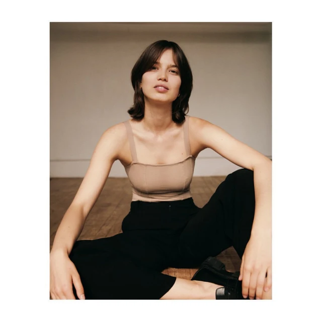 A young adult woman with short brunette hair sits on a wooden floor in a beige top and black trousers in a neutral indoor environment.