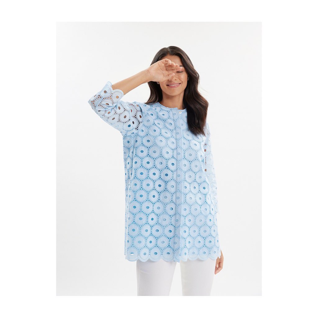 Fashionable adult female poses in an eye-catching baby blue tunic top with intricate circle cutouts, styled with white pants.