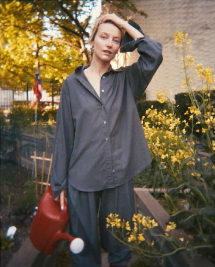Adult female in casual gray shirt and pants holding red watering can surrounded by yellow wildflowers in an outdoor setting.