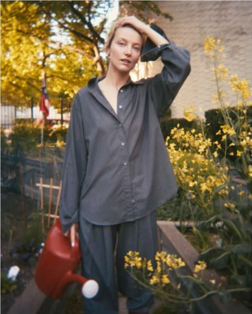 Adult female in casual gray shirt and pants holding red watering can surrounded by yellow wildflowers in an outdoor setting.