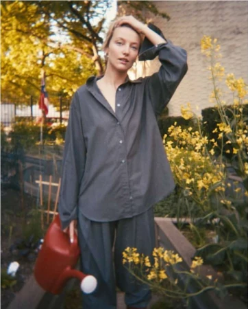 Adult female in casual gray shirt and pants holding red watering can surrounded by yellow wildflowers in an outdoor setting.