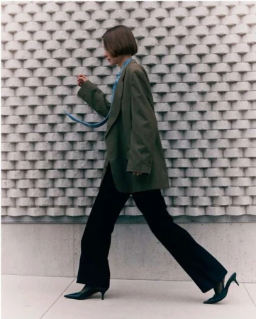 A woman in an oversized blazer and black pants strides confidently against a textured wall, showcasing a blend of modern fashion and urban style.