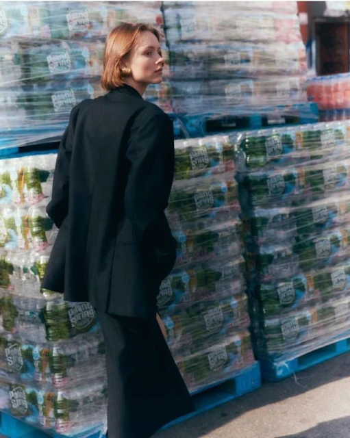 Woman in a black suit walking past pallets stacked with plastic-wrapped bottled water, side view, natural lighting, shallow depth of field.
