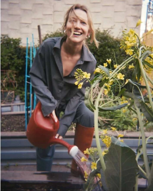 Blonde woman in grey overalls watering a garden with yellow flowers using a red watering can, smiling in an outdoor setting.