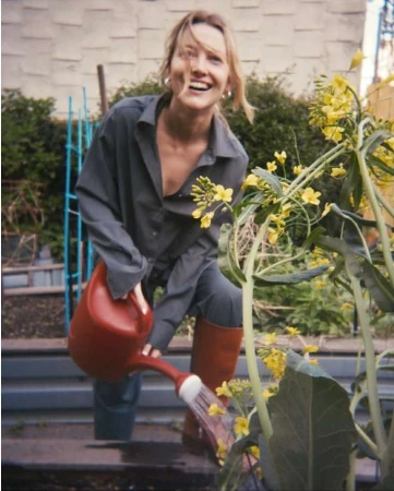 Blonde woman in grey overalls watering a garden with yellow flowers using a red watering can, smiling in an outdoor setting.