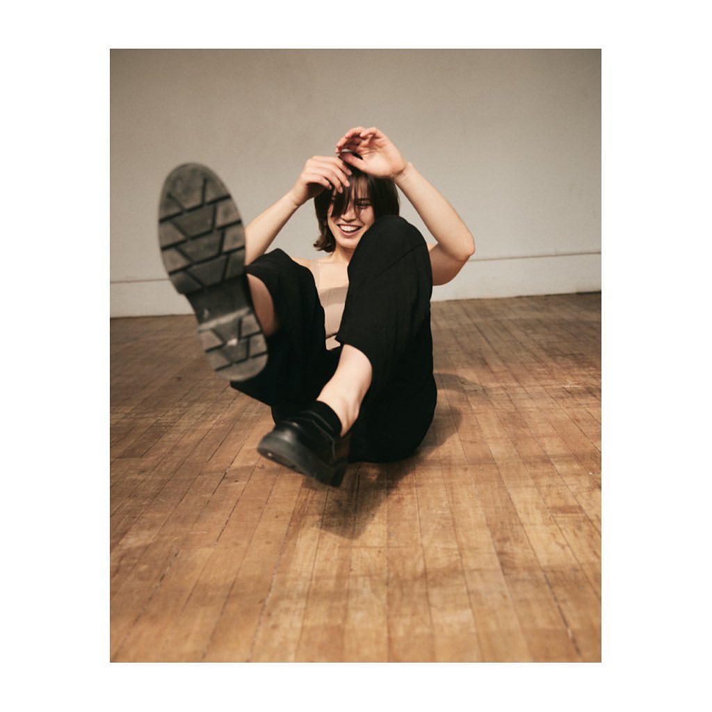 A smiling woman with dark hair sits on a wooden floor, wearing black pants and lifting a black shoe to the camera.