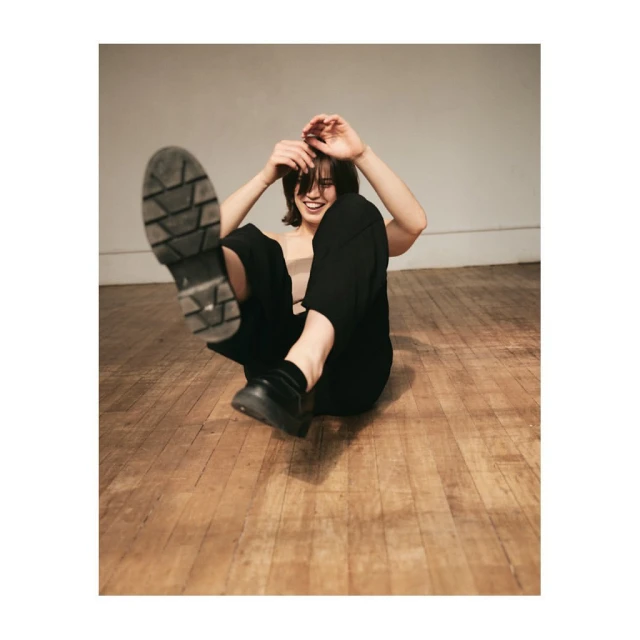 A smiling woman with dark hair sits on a wooden floor, wearing black pants and lifting a black shoe to the camera.