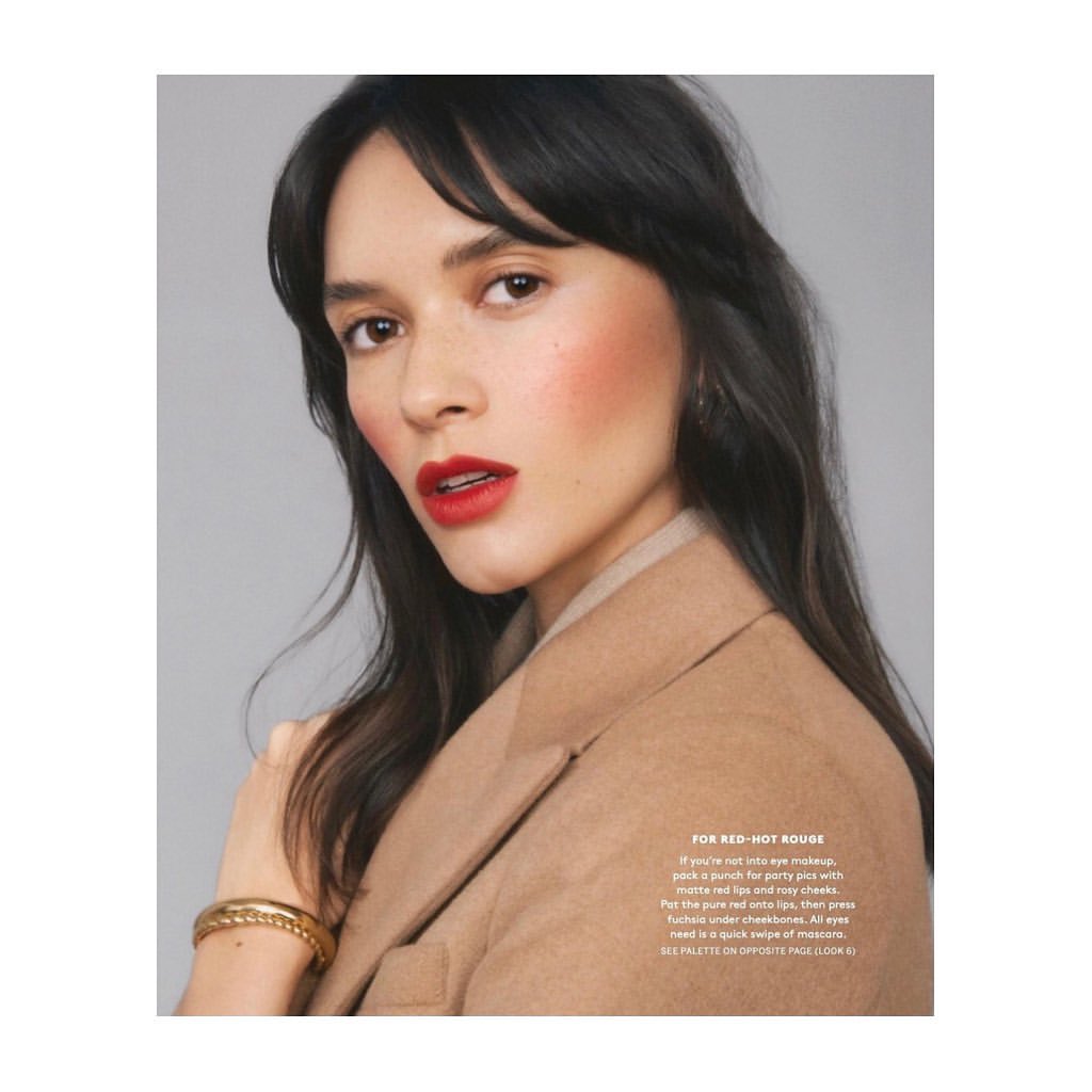Close-up of a woman with dark hair and red lipstick wearing a tan coat against a neutral grey background in a beauty editorial.
