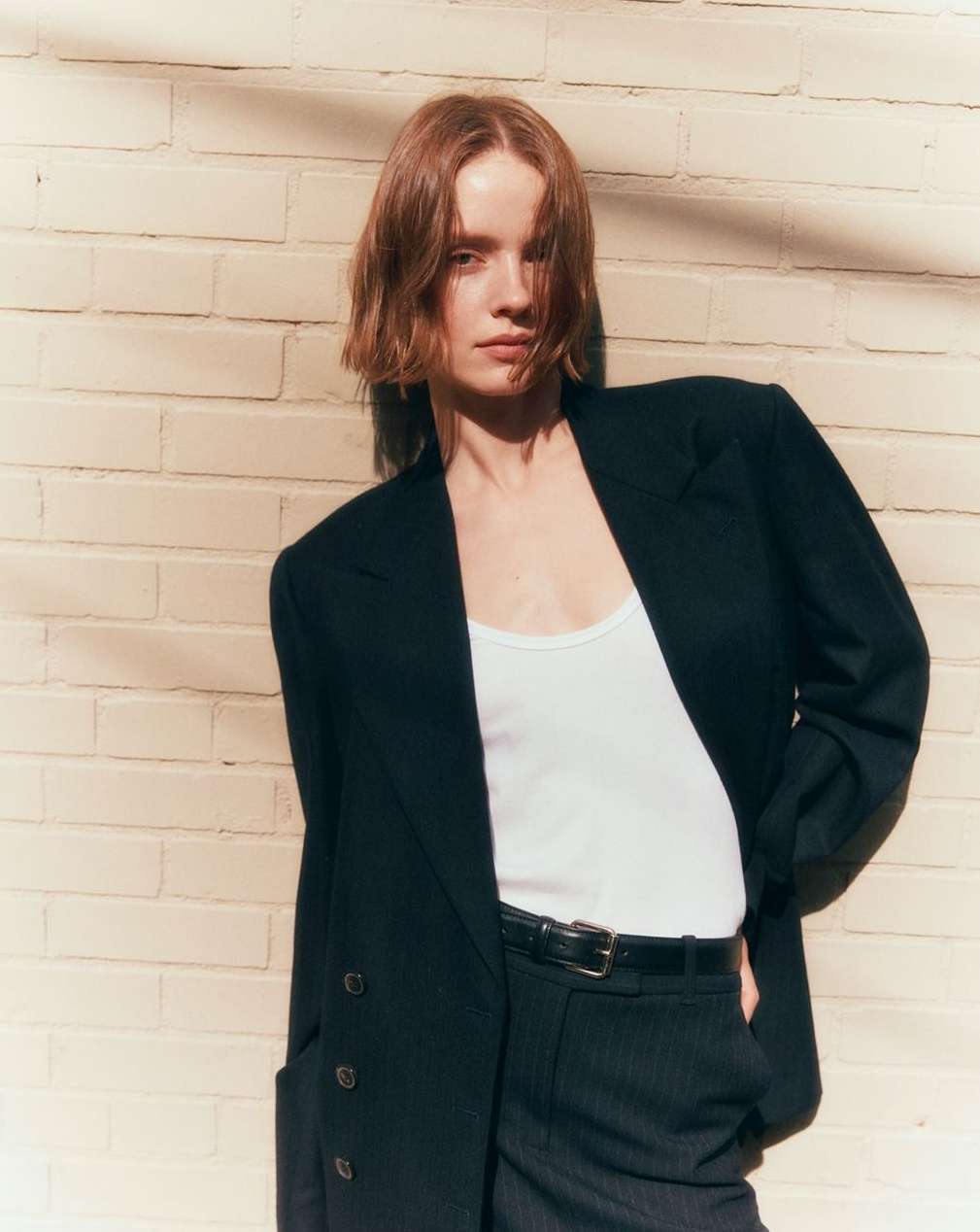 Fashion portrait of a red-haired woman in a black blazer and pinstripe pants, posing in front of a beige brick wall in warm sunlight.