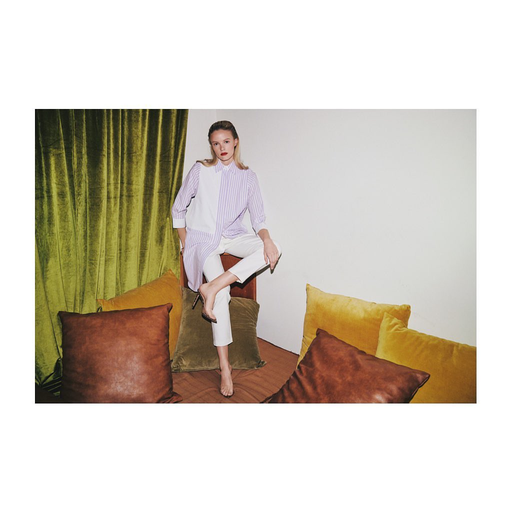 A slender woman poses indoors, wearing a striped top and white pants, perched atop a seat among assorted colored pillows, against a white wall.