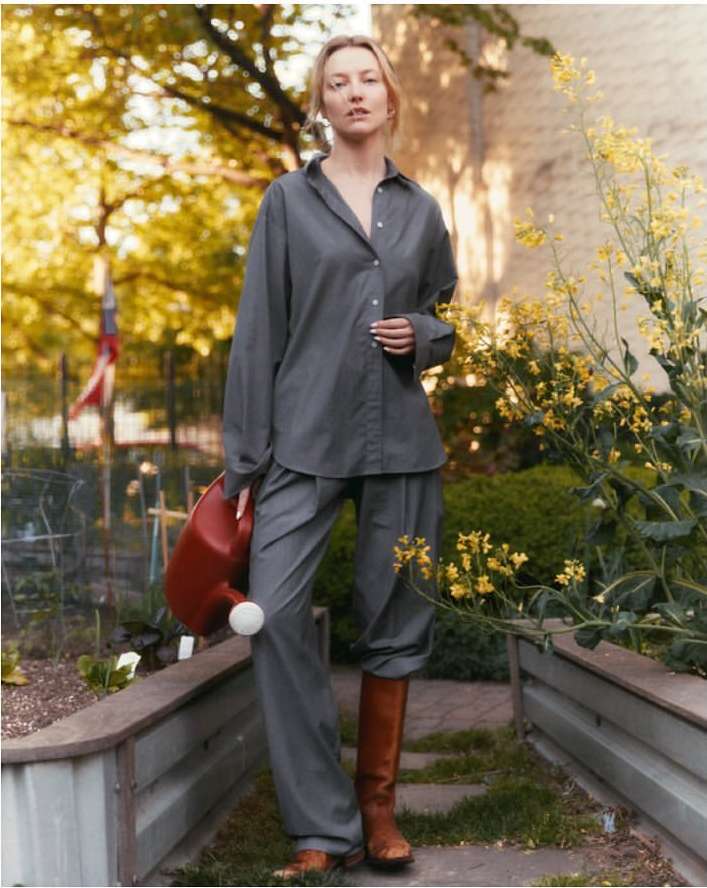 Woman in grey shirt and pants stands in a garden with raised beds, wearing boots and holding a red watering can in a bright outdoor setting.