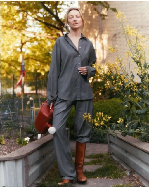 Woman in grey shirt and pants stands in a garden with raised beds, wearing boots and holding a red watering can in a bright outdoor setting.