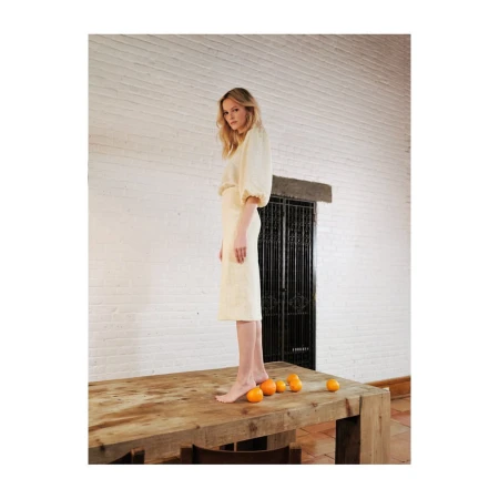 Woman in pale outfit standing on a wooden table among oranges against a white brick wall