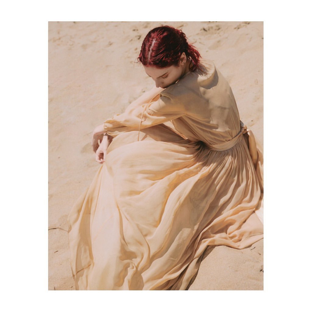 Woman in flowing beige dress seated on tan sand, looking down with red hair. Warm, soft daylight with shallow depth of field.