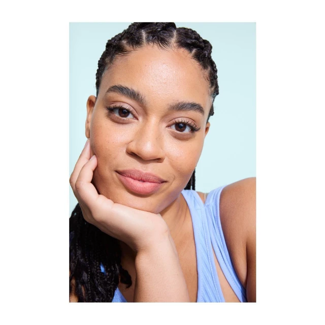 Close-up portrait of a smiling young woman with braided hair, wearing a blue tank top, posing against a pale green background.