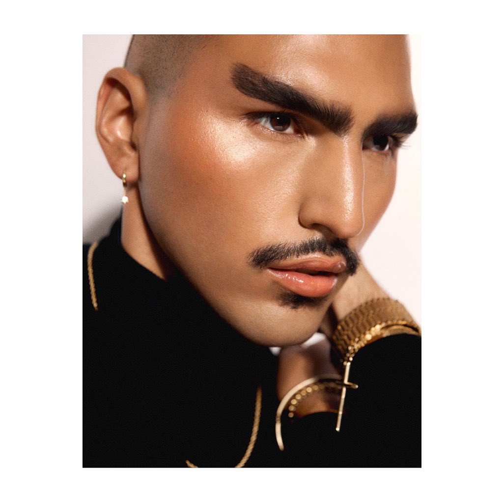 Close-up portrait of a person with a shaved head, thick styled eyebrows, and gold jewellery against a neutral background.