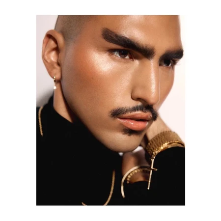Close-up portrait of a person with a shaved head, thick styled eyebrows, and gold jewellery against a neutral background.