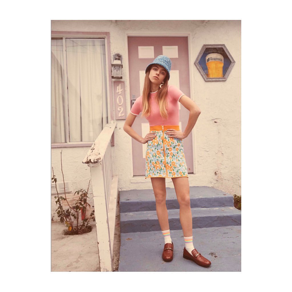 Woman posing in front of a building wearing a pink top, floral mini skirt, blue hat, and brown loafers with striped socks.