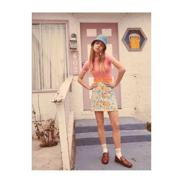 Woman posing in front of a building wearing a pink top, floral mini skirt, blue hat, and brown loafers with striped socks.