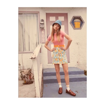 Woman posing in front of a building wearing a pink top, floral mini skirt, blue hat, and brown loafers with striped socks.