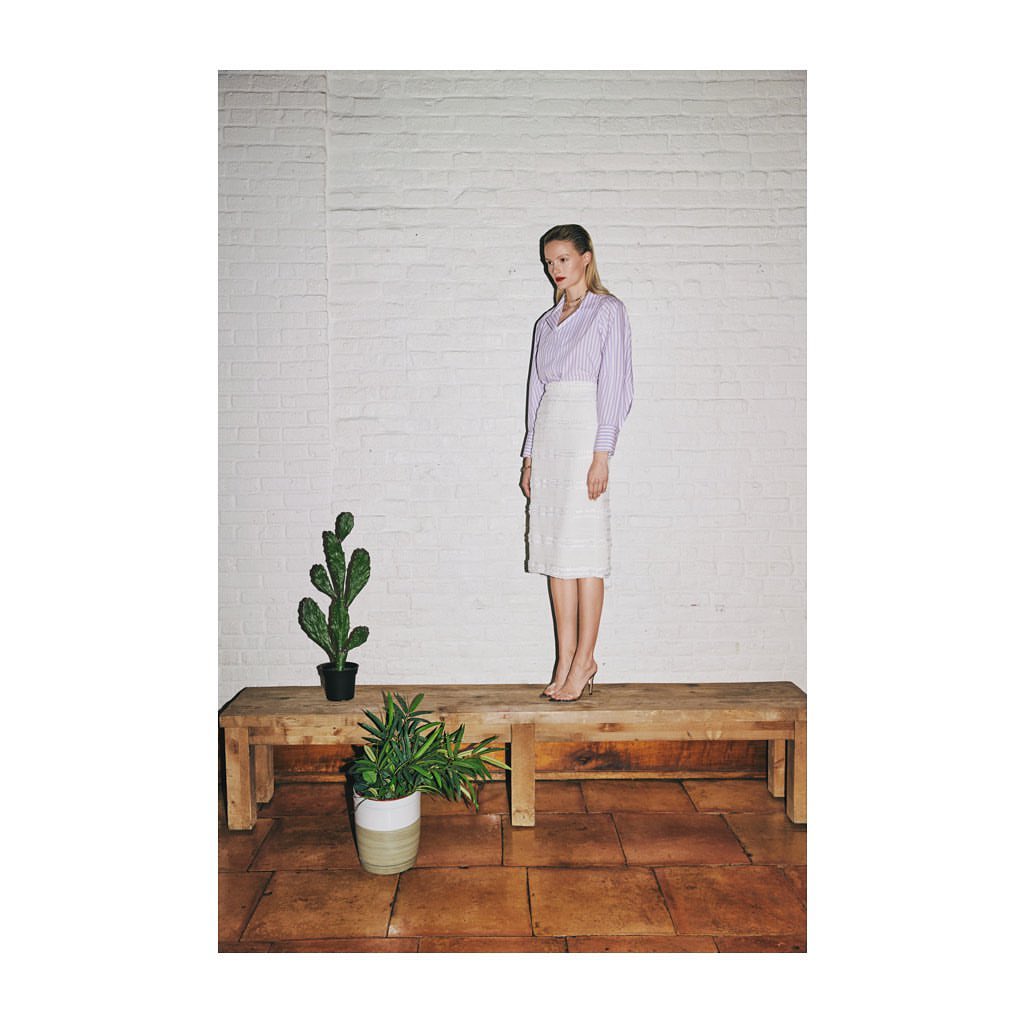 Adult woman poses on wooden platform wearing skirt and blouse in front of brick wall near potted cactus and house plant indoors.