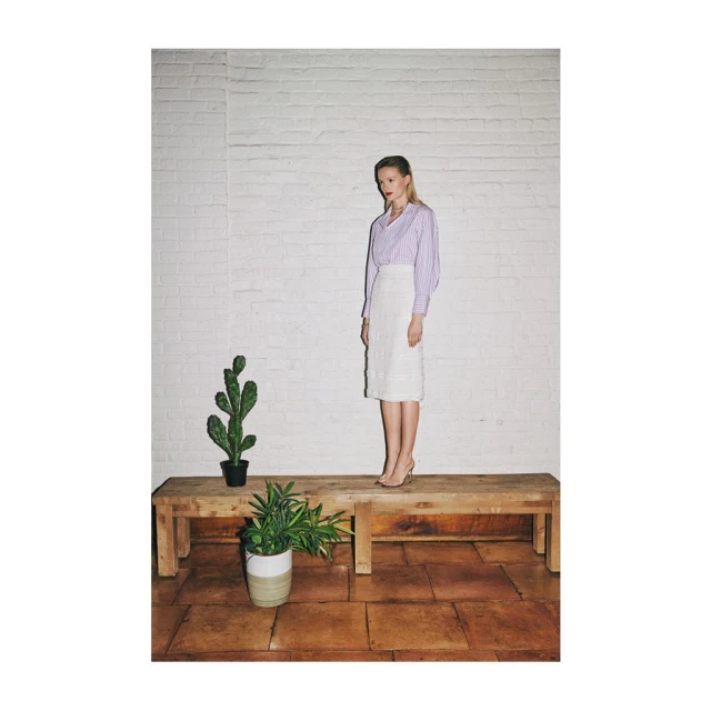 Adult woman poses on wooden platform wearing skirt and blouse in front of brick wall near potted cactus and house plant indoors.
