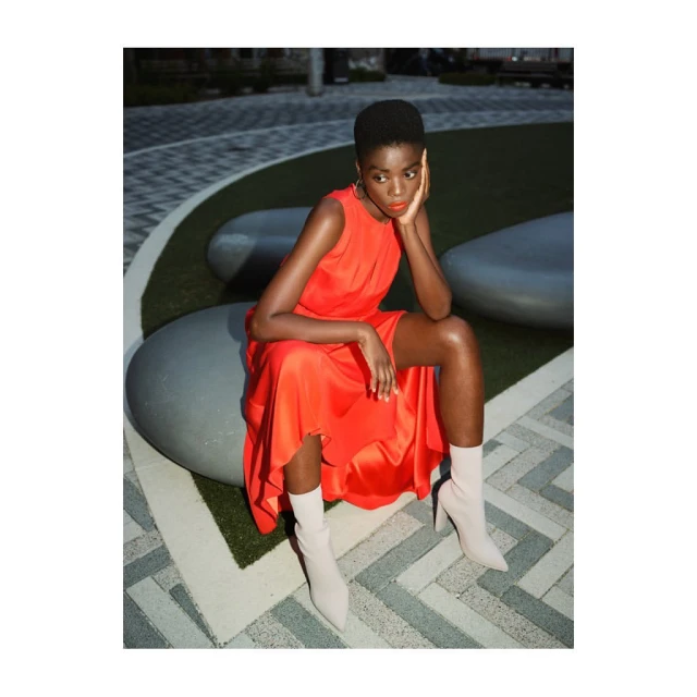 Attractive woman sits on an outdoor bench in a red dress and light-colored heeled boots, posing thoughtfully on a patterned brick surface.