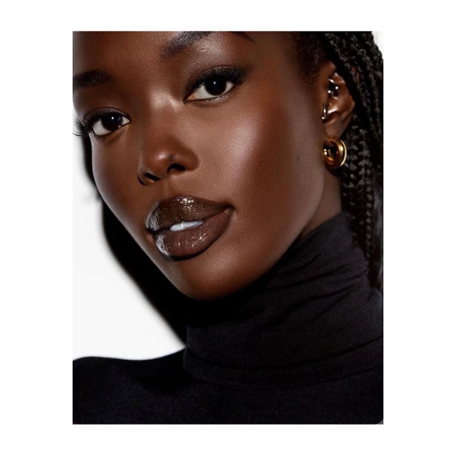 Close-up portrait of a young Black woman with braided hair, shiny brown lipstick, wearing a black turtleneck against a white background.