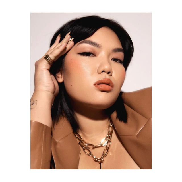 Portrait of an Asian woman with dark hair and natural makeup wearing a tan jacket and gold necklace against a white background.