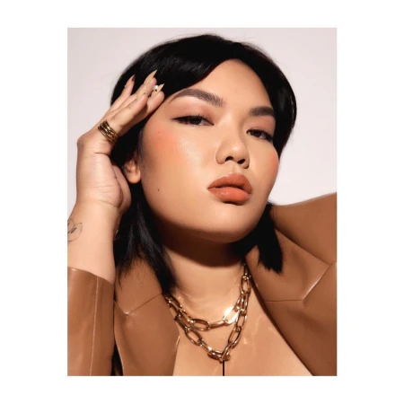 Portrait of an Asian woman with dark hair and natural makeup wearing a tan jacket and gold necklace against a white background.