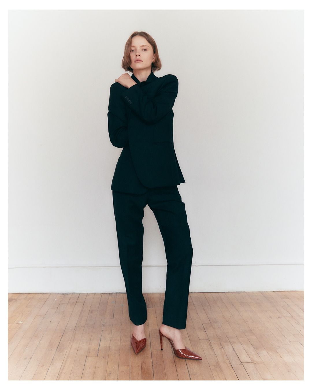 A stylish woman in a dark suit poses against a white wall on a wood floor, accented by brown heels, for a fashion editorial.